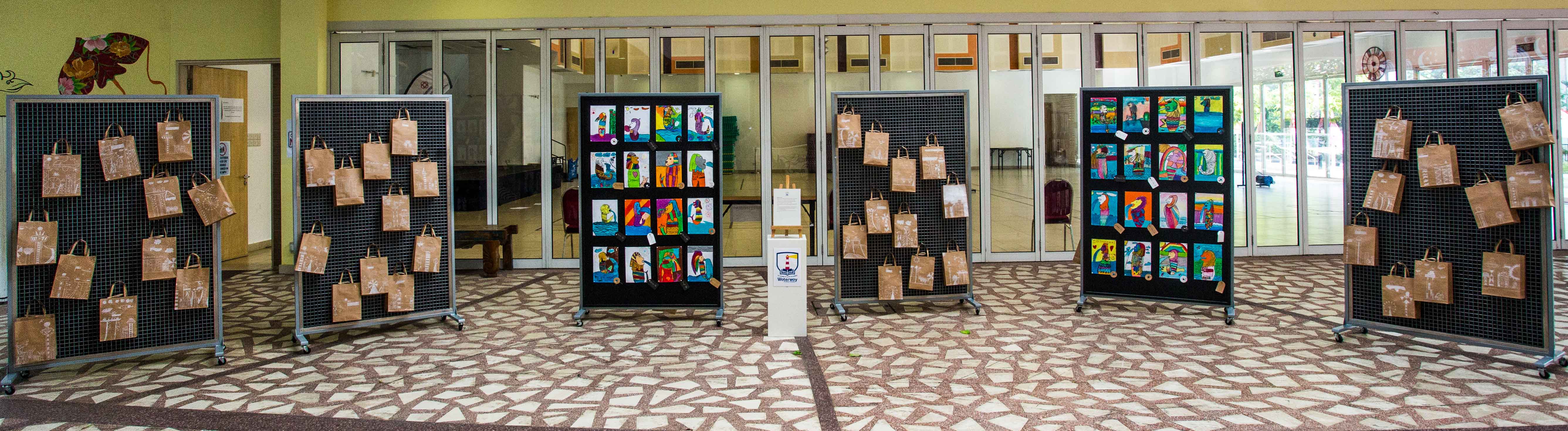 Several black display panels in a lobby holding colorful artworks and brown paper bags in an indoor exhibition.